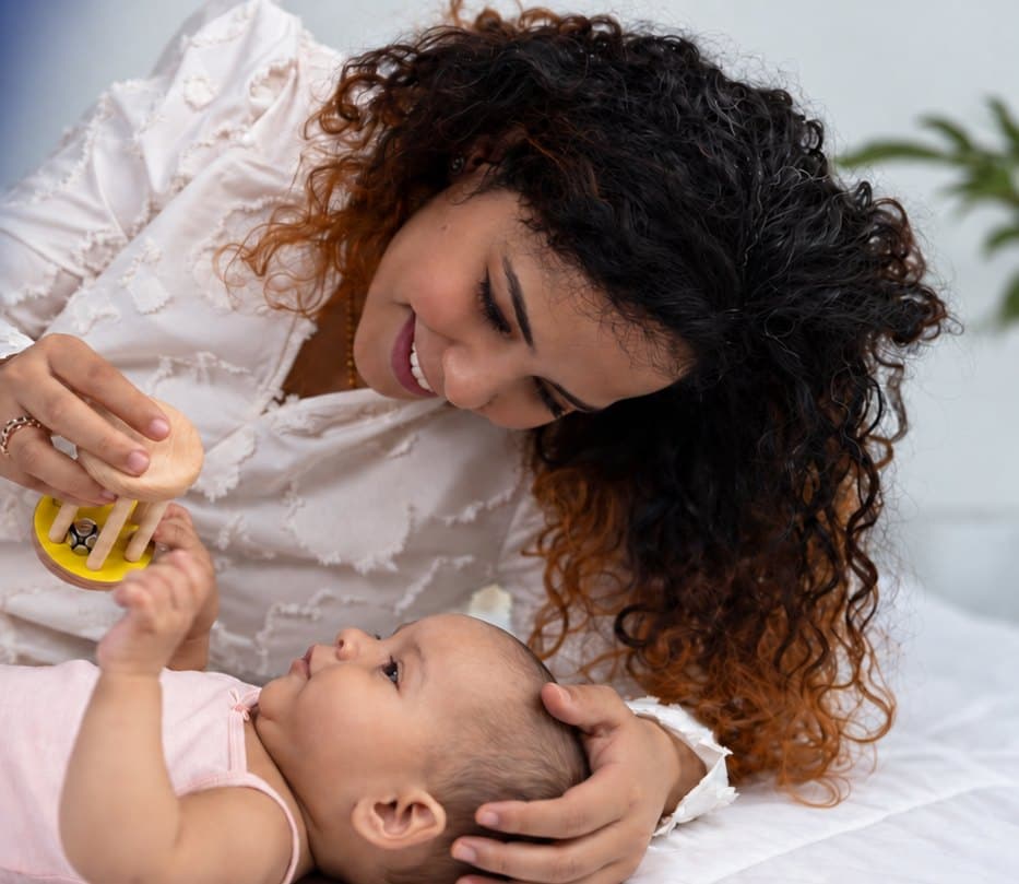A mother holds a wooden rattle above her newborn, who reaches up for it
