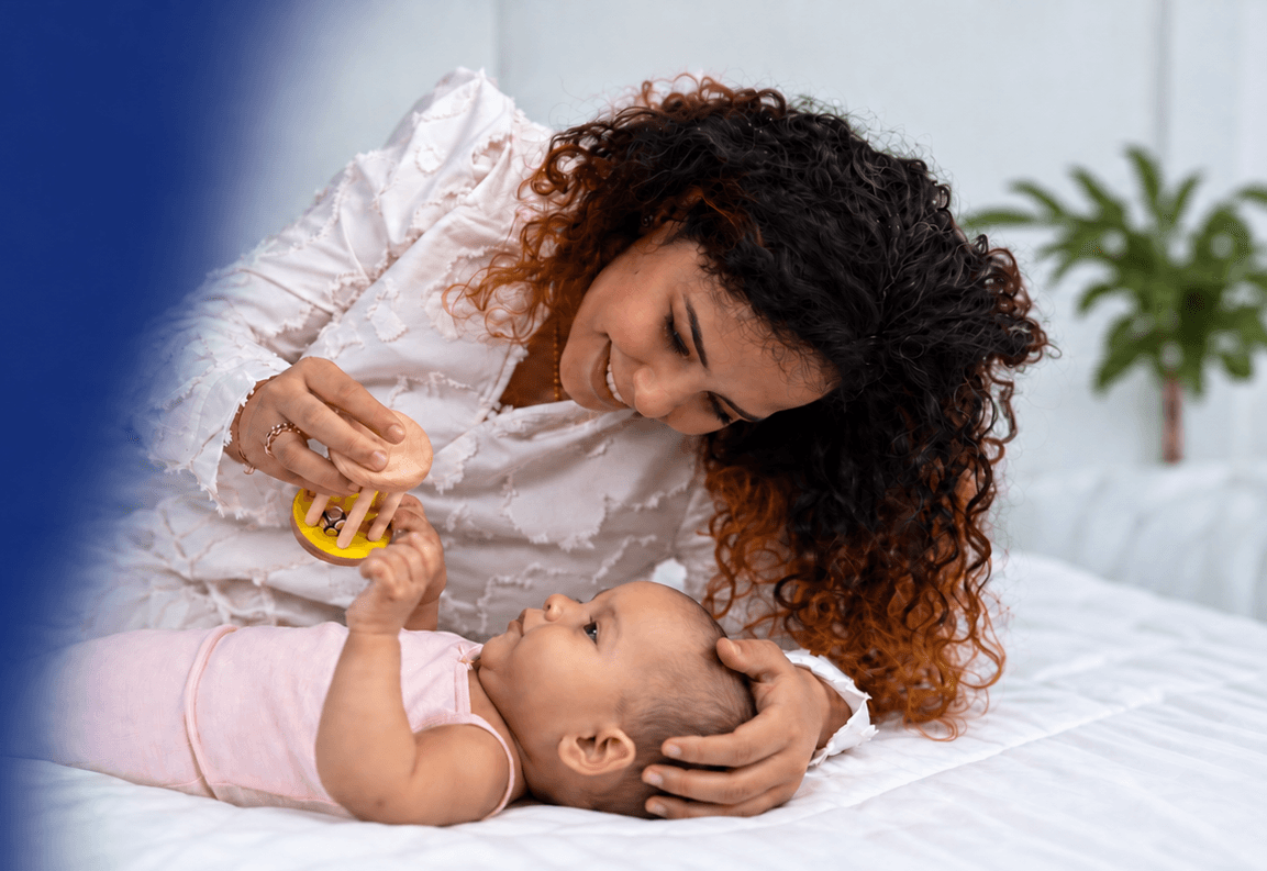 A mother holds a wooden rattle above her newborn, who reaches up for it.