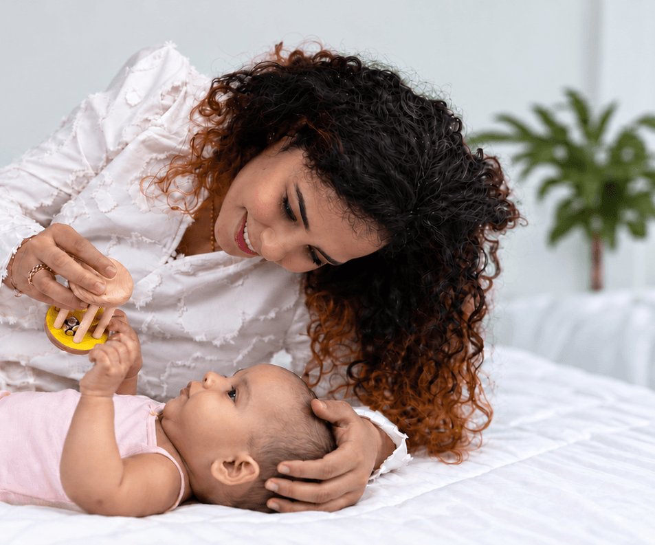 A mother holds a wooden rattle above her newborn, who reaches up for it.
