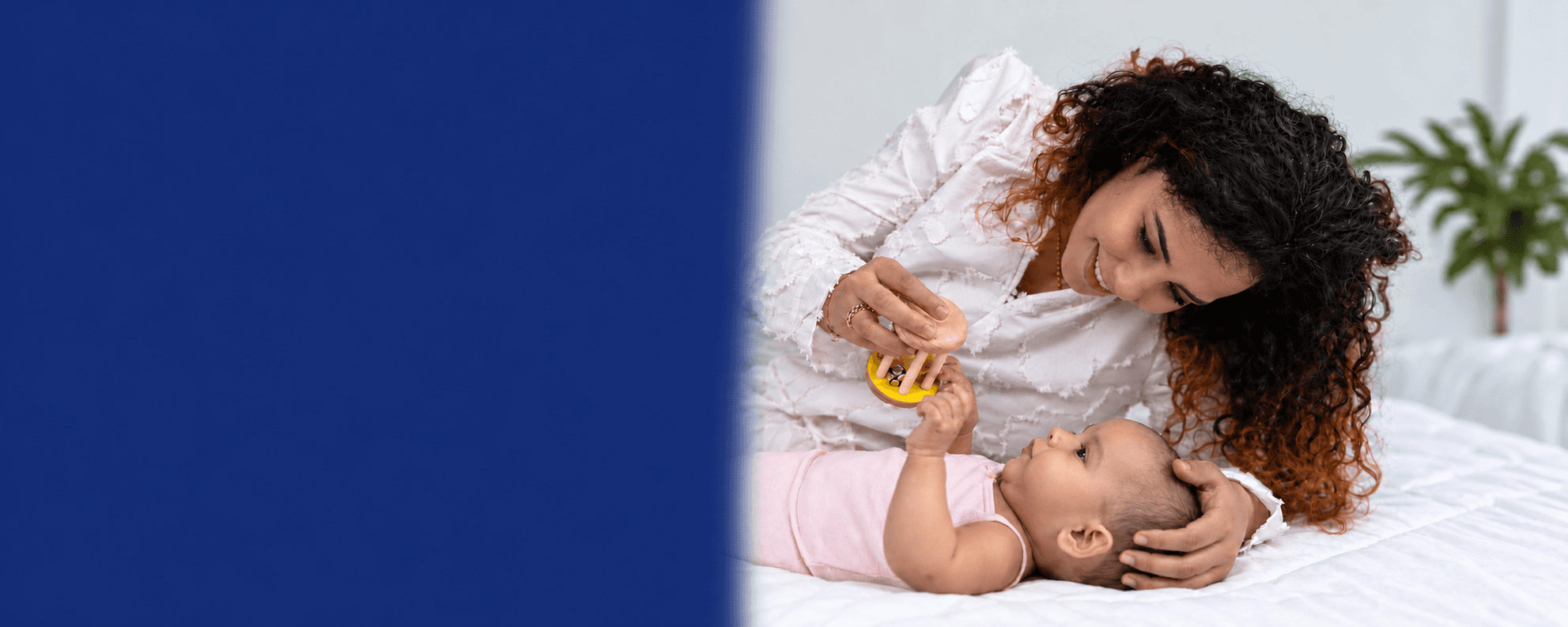 A mother holds a wooden rattle above her newborn, who reaches up for it.